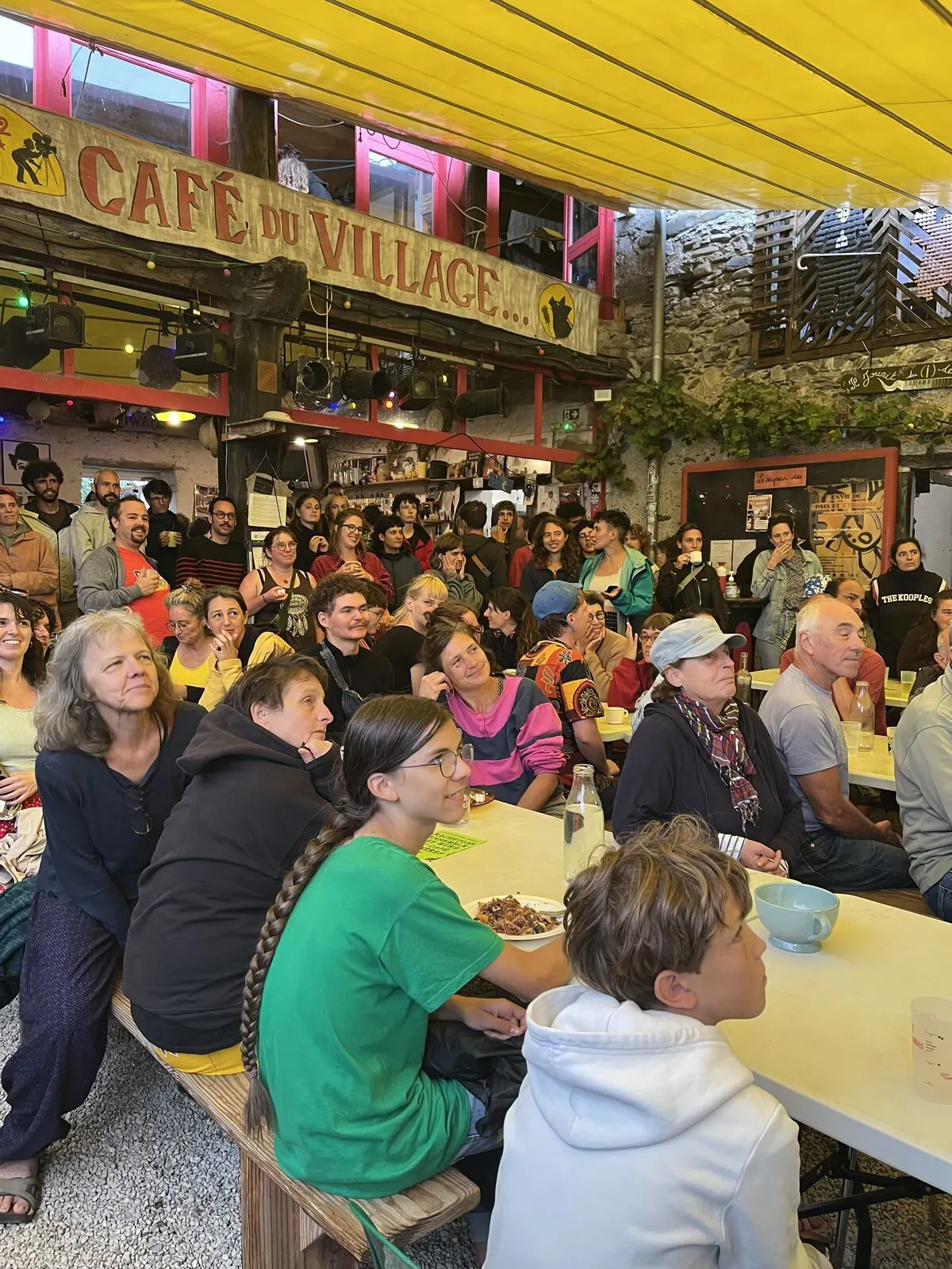 La cour du Café pleine de monde, sous l’enseigne Café du Village